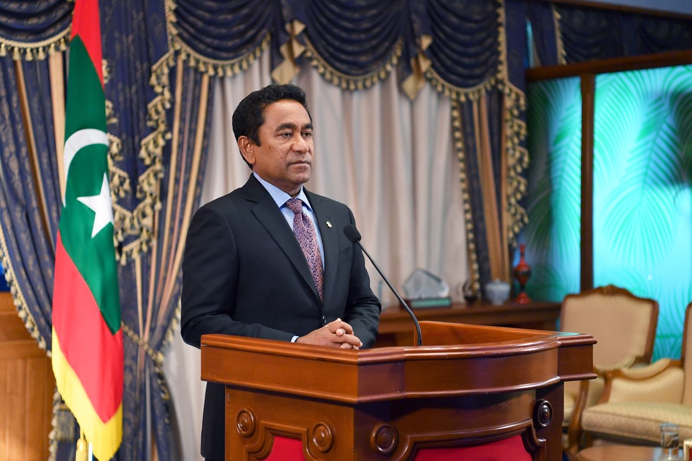 ޢަބްދުﷲ ޔާމީން speaking at a podium beside the national flag of the Maldives.